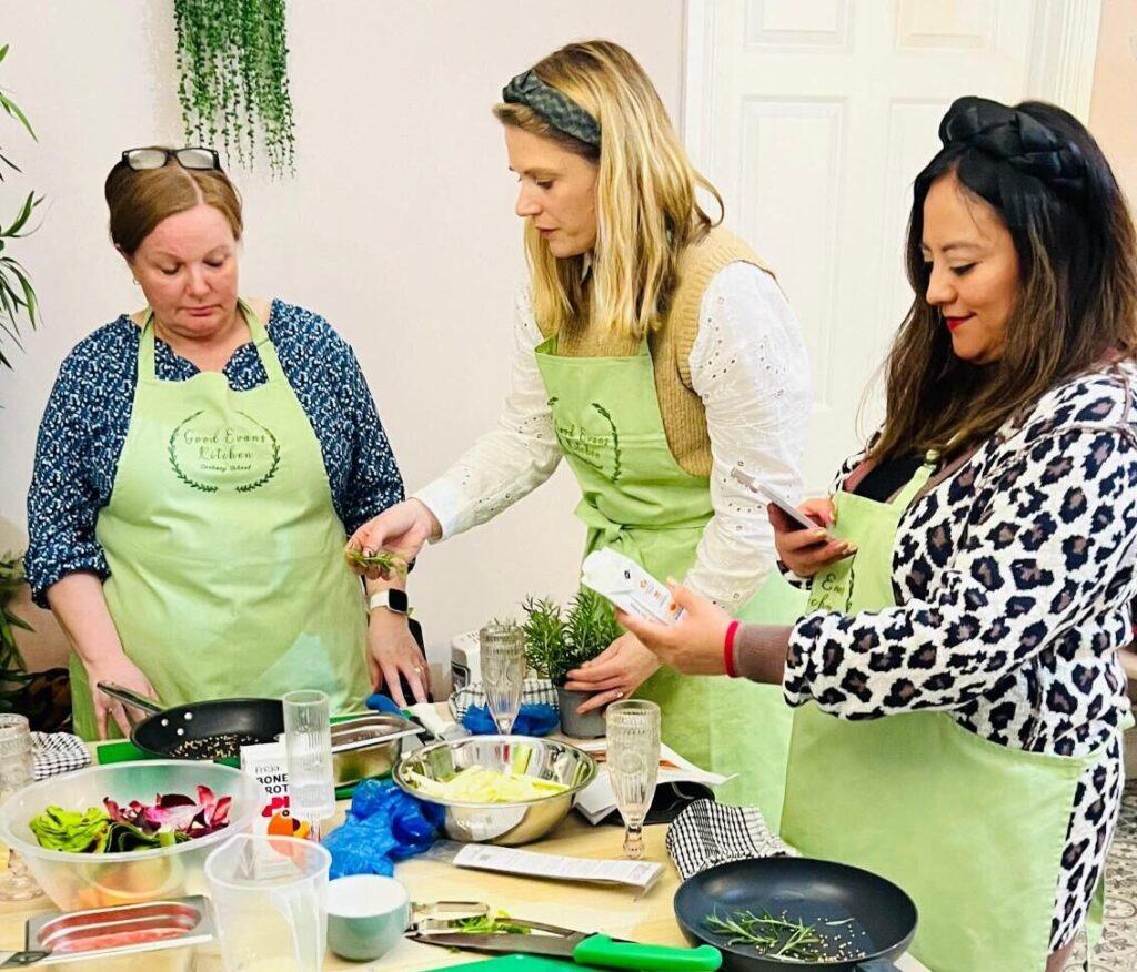 Three women in green Good Evans Kitchen aprons cooking together at a workshop