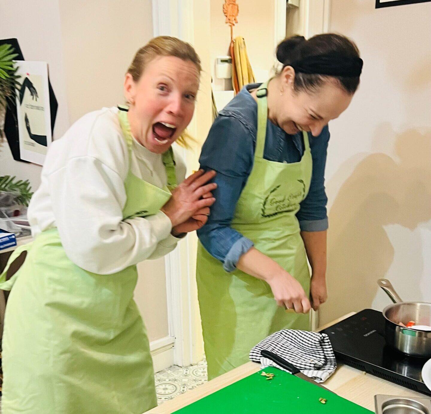 Two women laughing while cooking in green aprons